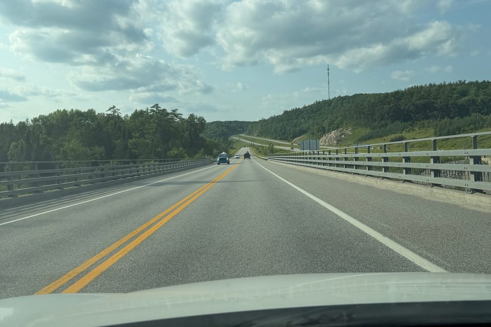 An open two-lane road stretching through forest beneath a cloud-filled sky.