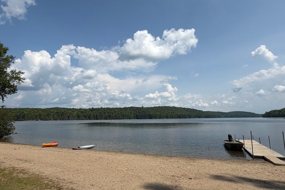 A quiet lake with a sandy beach, dock, kayaks, and a forested shoreline.