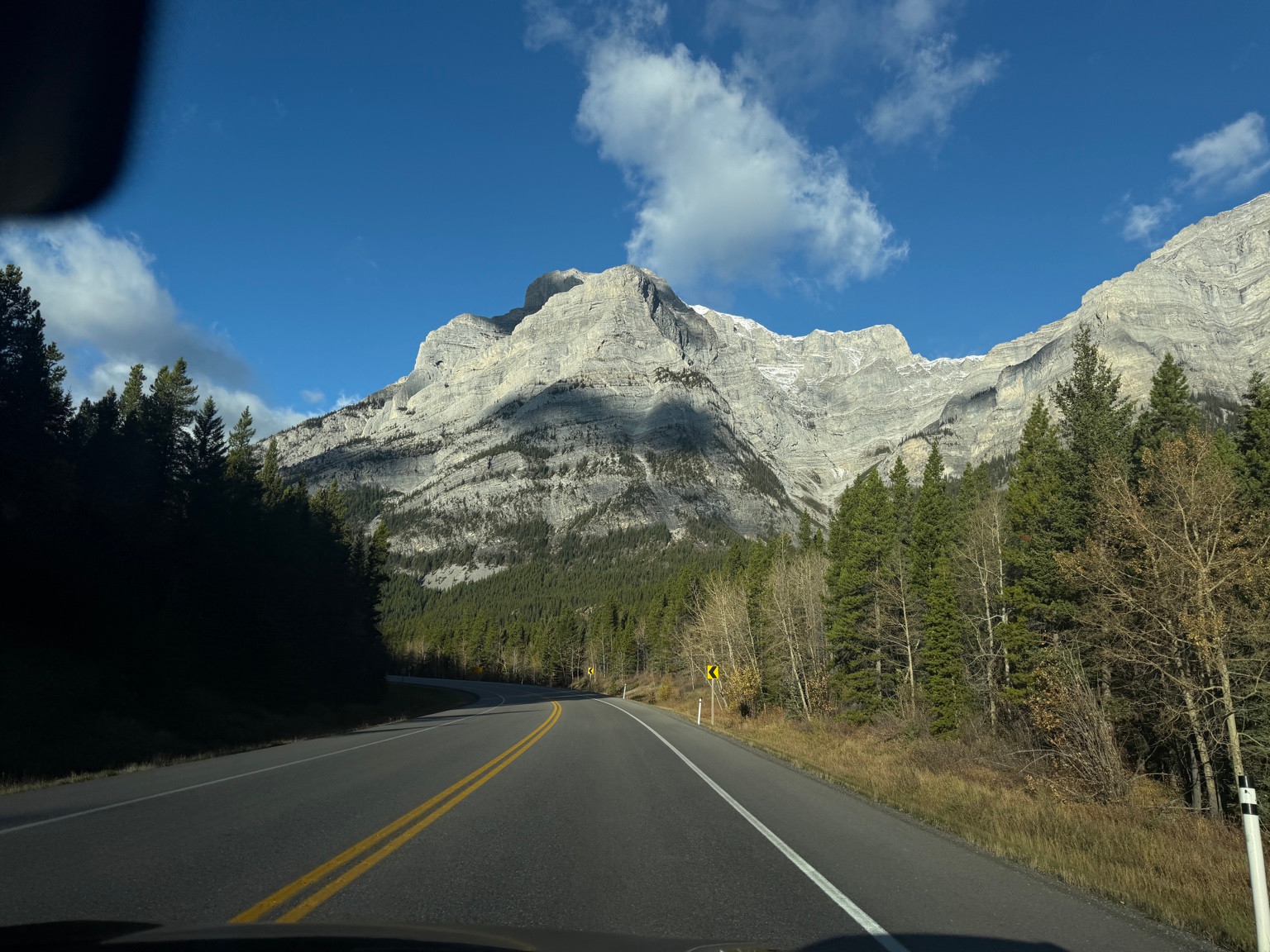 A curving two-lane road passes through evergreen trees below a rocky mountain ridge.
